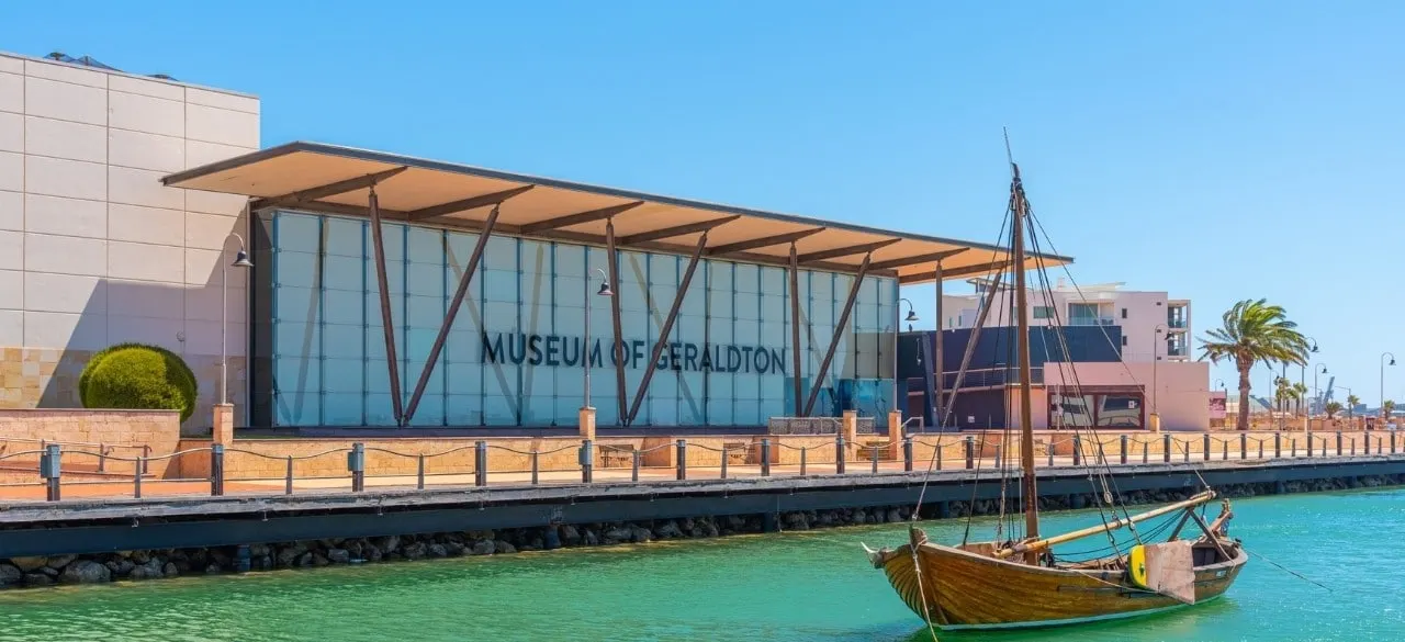 The modern glass-fronted Museum of Geraldton across a turquoise waterway with a wooden sailboat