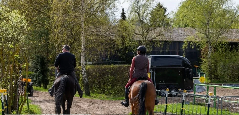 Two riders on horseback following a dirt path past a horse float and green trees